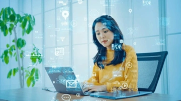 A woman in a yellow sweater working on a laptop with digital icons floating around her, indicating data and technology.